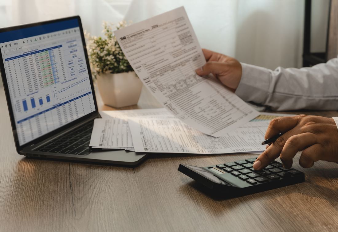 Hands working a calculator beside printed statements and a laptop